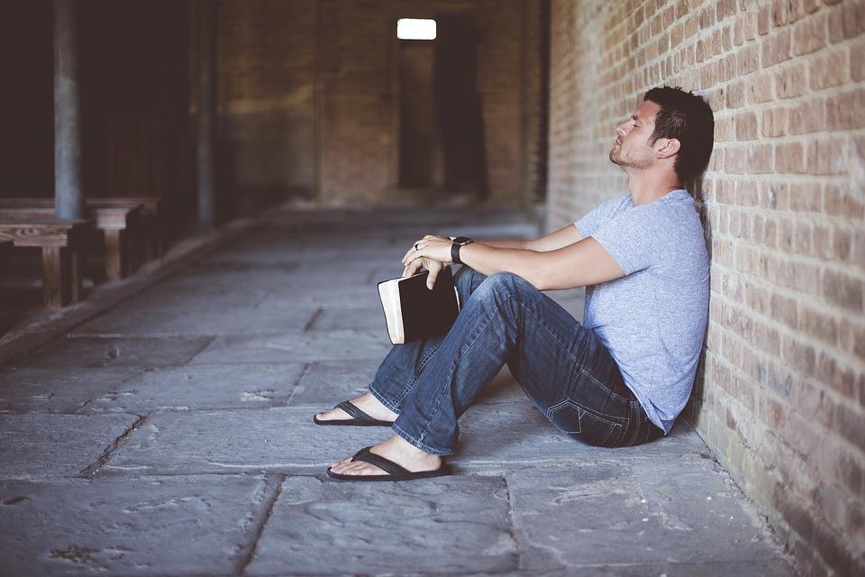 mies istuu kivilattialla nojaten tiiliseinään, kädessään Raamattu